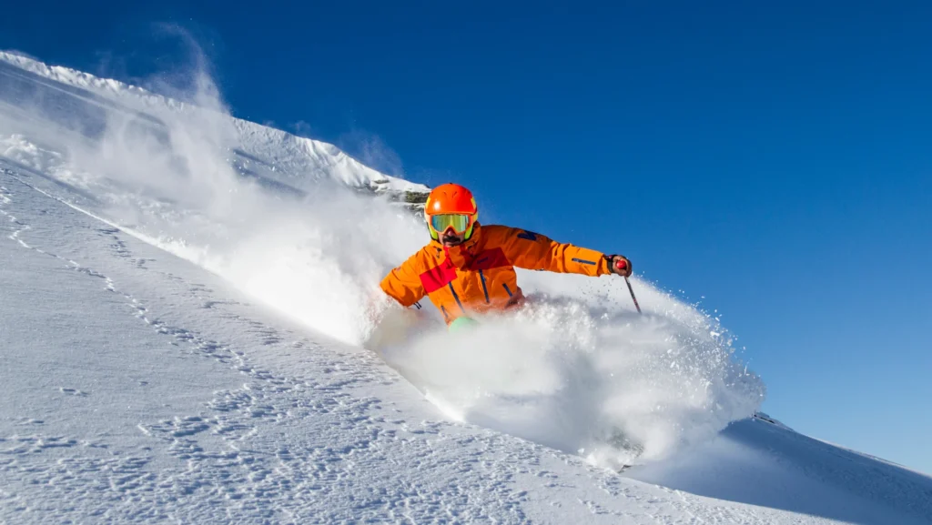 A skier in an orange jacket and helmet carving through fresh powder on a steep slope under a clear blue sky. The dynamic movement and snow spray capture the thrill of high-altitude skiing.