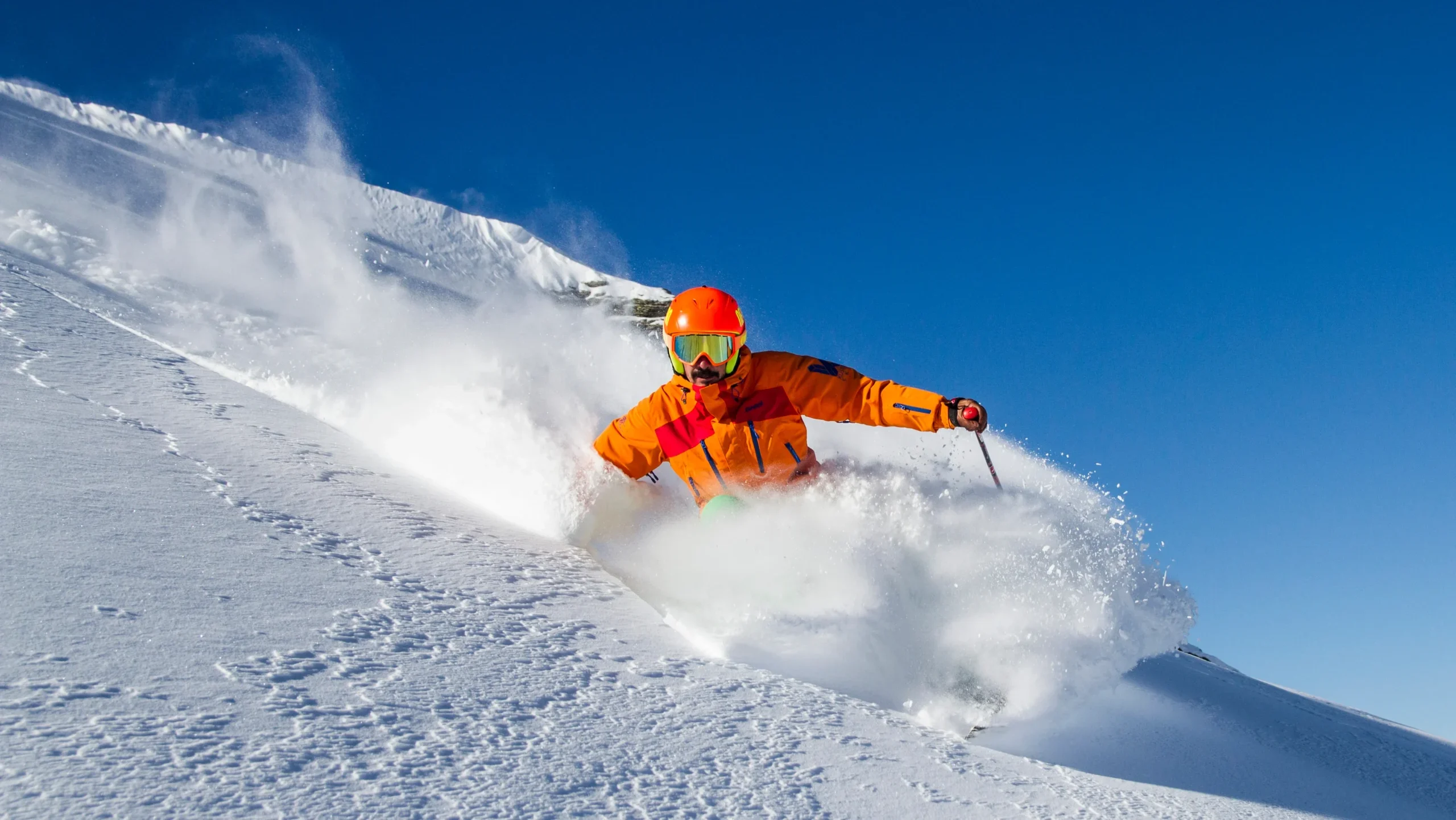 A skier in an orange jacket and helmet carving through fresh powder on a steep slope under a clear blue sky. The dynamic movement and snow spray capture the thrill of high-altitude skiing.