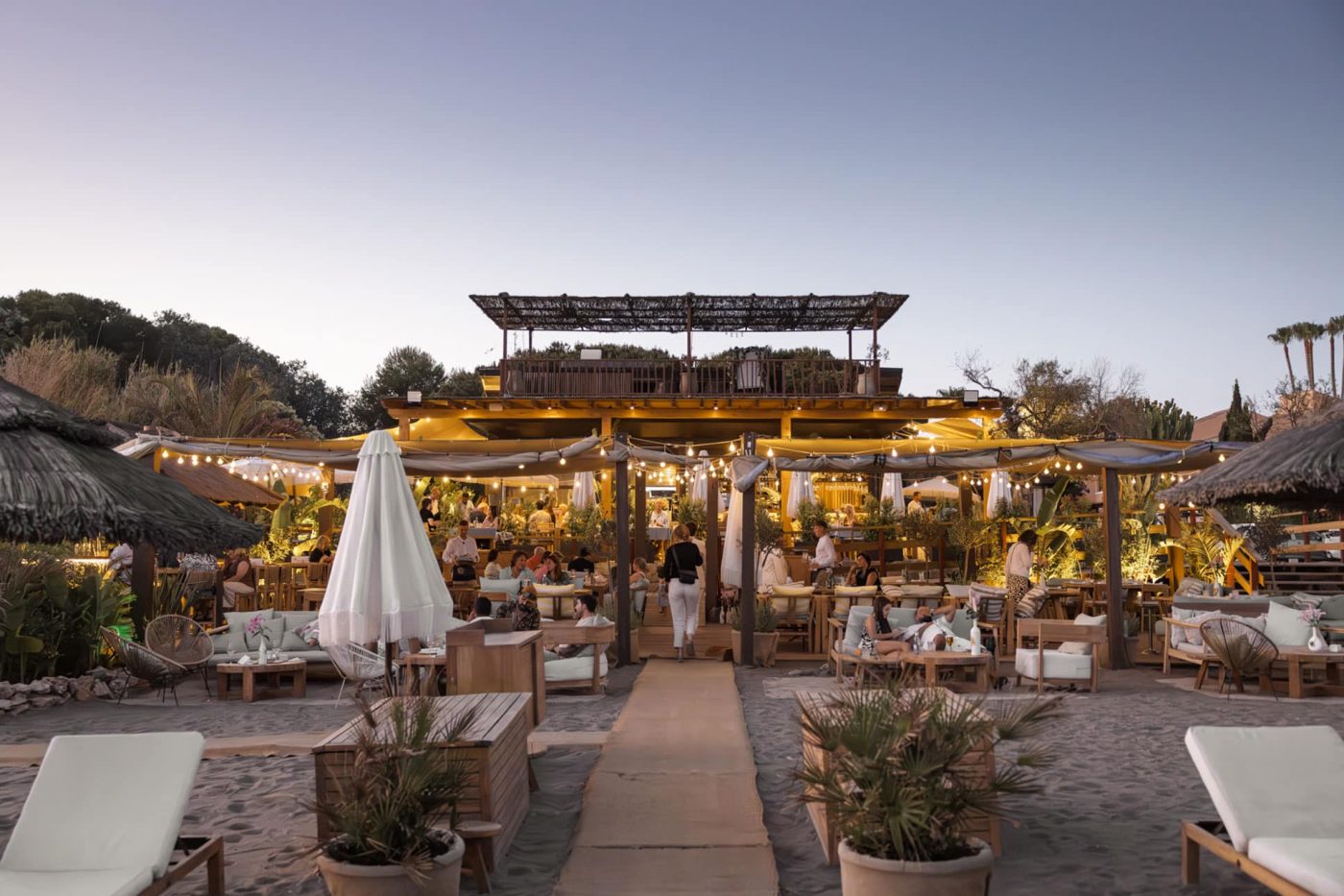Estepona Beach Clubs. Salduna Beach terrace in Estepona at dusk, wooden pergola and string lights over beachfront seating on the sand
